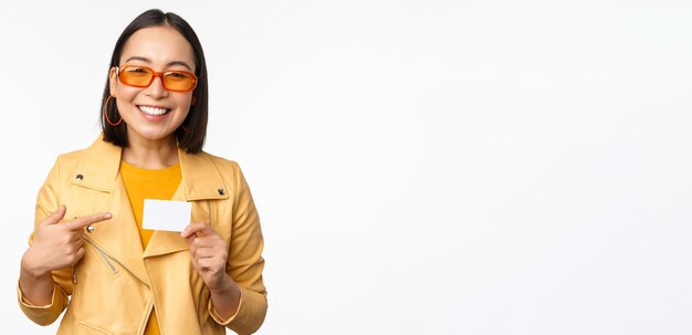 Portrait of beautiful modern asian girl in sunglasses smiling happy showing credit card standing over white background