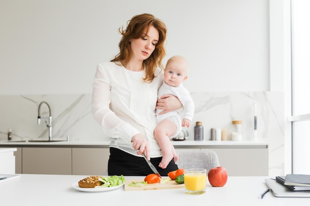 Портрет красивой матери, держащей своего милого маленького ребенка, стоя и готовя на кухне
