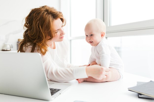 아름다운 어머니가 앉아 있고 근처에 노트북이 있는 행복한 어린 아기를 꿈꾸며 바라보는 초상화