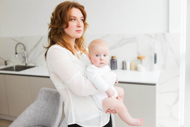 Ritratto di bella madre in piedi e sognante che guarda da parte mentre tiene in mano il suo piccolo bambino carino