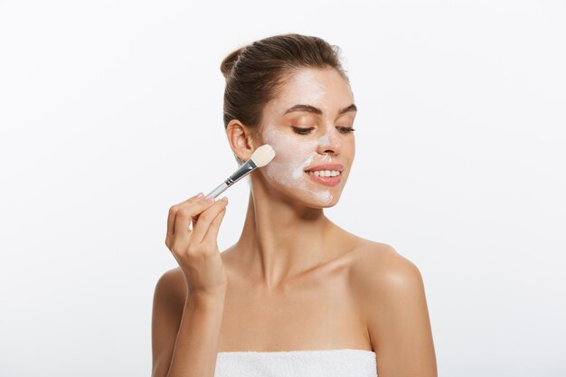 Portrait beautiful naked woman apply cosmetic white clay mask on with brush isolated on a white back