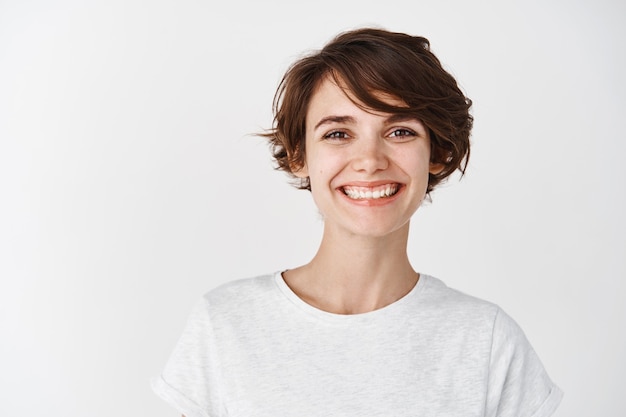 Ritratto di bella ragazza naturale senza trucco, sorridente felice, in piedi in maglietta contro il muro bianco