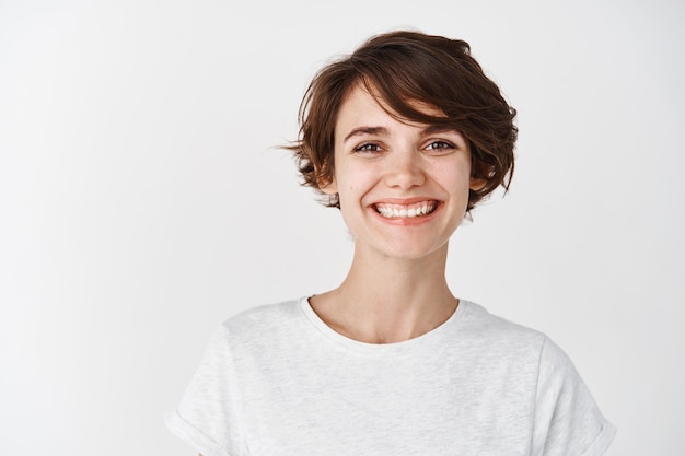 Портрет красивой естественной девушки без макияжа, счастливой улыбкой, стоящей в футболке у белой стены