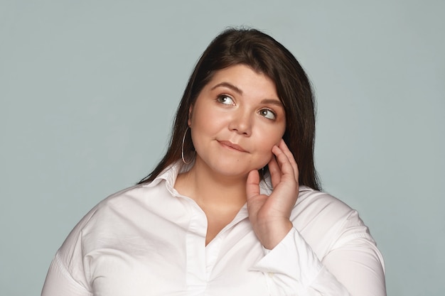 Portrait of beautiful overweight young female employee wearing white shirt and round earrings looking up and biting lip, having pensive expression, thinking what healthy food eat for lunch