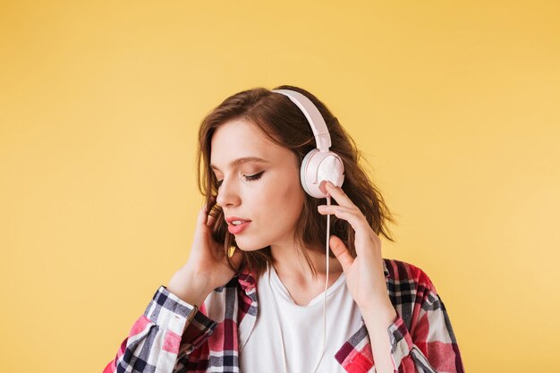 ピンクの背景の上に立ってヘッドフォンで音楽を聞いているカラフルなシャツの美しい物思いにふける女性の肖像画