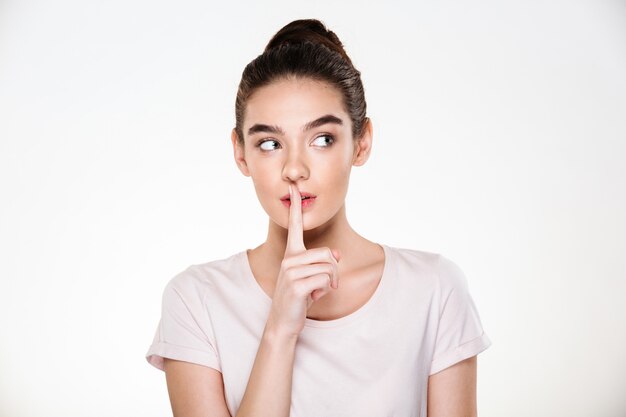 Portrait of beautiful pensive young woman with perfect skin holding index finger on lips asking to keep silence or secret