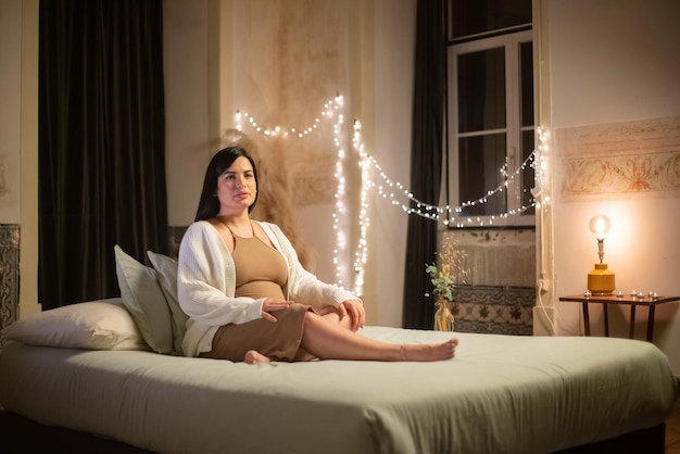 ベッドの上の美しい妊婦の肖像画。カメラを見ているドレスとカーディガンの裸足の女性。妊娠、期待の概念