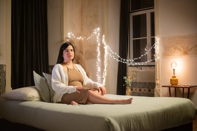 Portrait of beautiful pregnant woman on bed. Barefoot woman in dress and cardigan looking at camera. Pregnancy, expectation concept