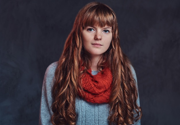 따뜻한 스웨터와 스카프를 두른 아름다운 빨간 머리 소녀의 초상화는 어두운 질감의 배경에서 격리된 카메라를 바라보고 있습니다.