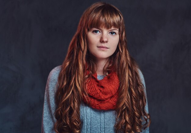 Portrait of a beautiful redhead girl wearing a warm sweater and scarf looking at a camera, isolated on a dark textured background.