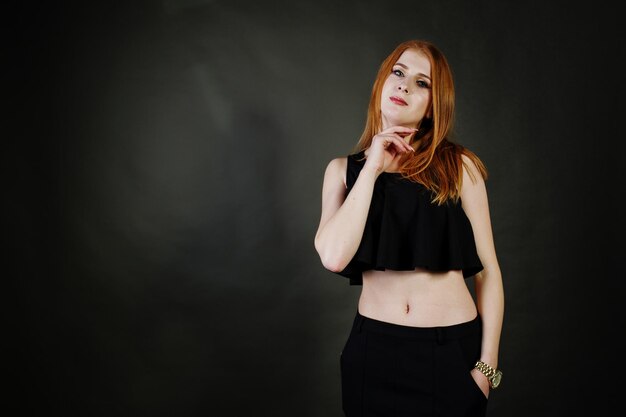 Portrait of a beautiful redheaded girl in black top and black skirt posing in the studio next to the grey wall