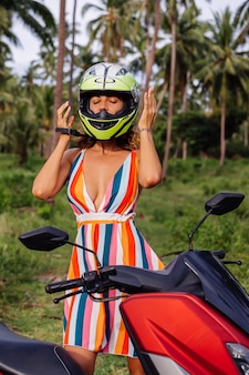 Ritratto di donna bella pilota in casco da motociclista verde giallo e vestito estivo leggero colorato nella giungla sul campo tropicale sotto le palme.