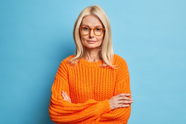 Portrait of beautiful serious middle aged woman keeps hands crossed wears spectacles and orange sweater looks confidently listens attentively interlocutor.