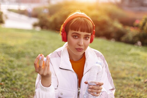 Portrait of beautiful shorthaired woman in white jacket holding water bottle Pretty girl sits on grass and listens to music in headphones