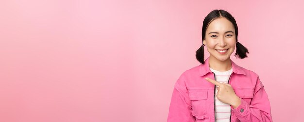 ピンクの背景の上に立っている広告バナープロモーションを示す左指を指している美しい笑顔のアジアの女の子の肖像画