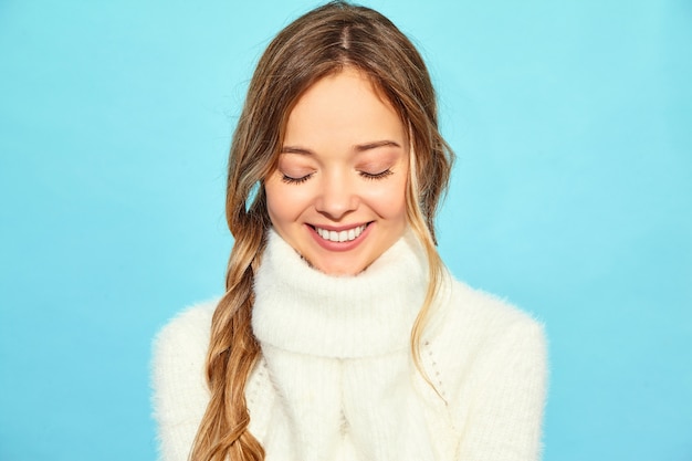 Free photo portrait of beautiful smiling blond gorgeous woman. woman standing in stylish white sweater, on blue wall. concept of winter