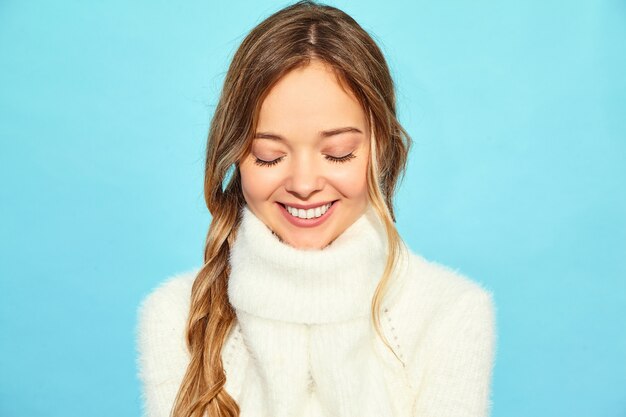 美しい笑顔金髪ゴージャスな女性の肖像画。青い壁に、スタイリッシュな白いセーターで立っている女性。冬の概念