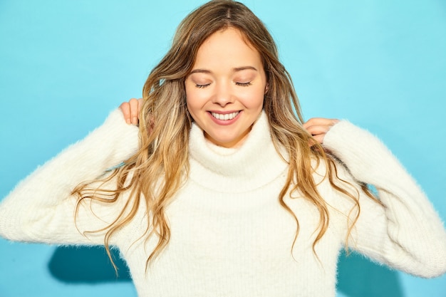 美しい笑顔金髪ゴージャスな女性の肖像画。青い壁に、スタイリッシュな白いセーターで立っている女性。冬の概念