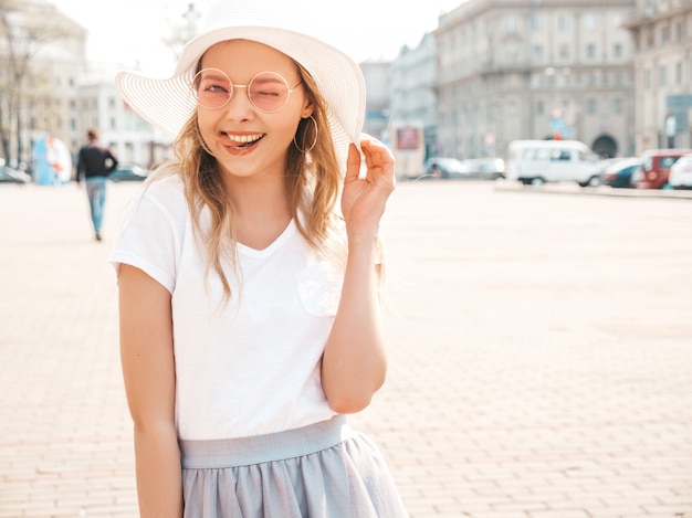 Il ritratto di bello modello biondo sorridente si è vestito in vestiti dei pantaloni a vita bassa dell'estate. ragazza d'avanguardia che propone in strada in occhiali da sole rotondi. donna divertente e positiva divertendosi in cappello