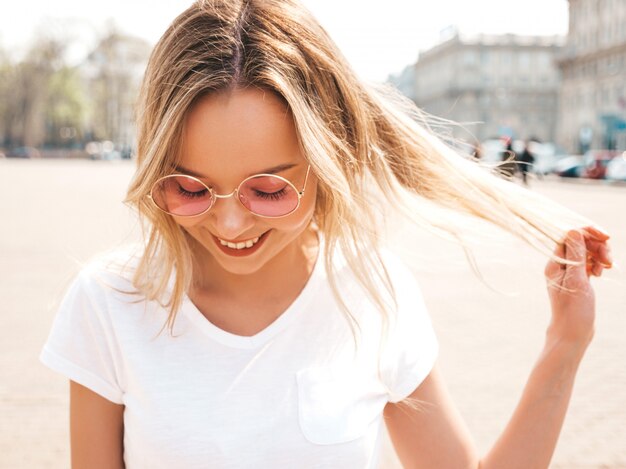 Portrait of beautiful smiling blond model dressed in summer hipster  clothes. Trendy girl posing in the street   in round sunglasses. Funny and positive woman having fun