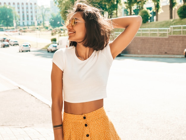 Foto gratuita il ritratto di bello modello castana sorridente si è vestito in vestiti dei pantaloni a vita bassa dell'estate. ragazza d'avanguardia che posa nei precedenti della via. divertimento donna divertente e positiva