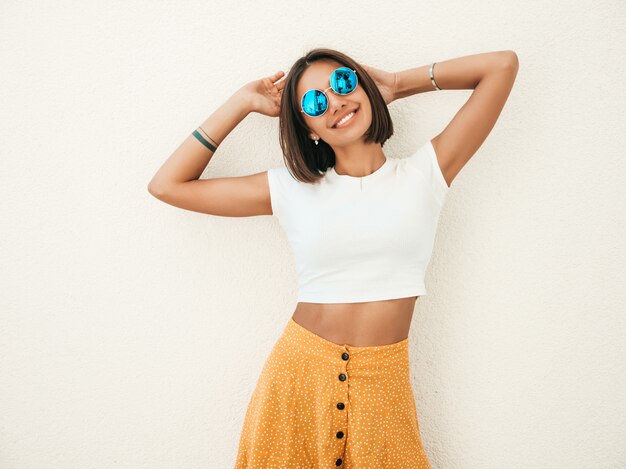 Portrait of beautiful smiling brunette model dressed in summer hipster  clothes. Trendy girl posing in the street near white wall. Funny and positive woman having fun