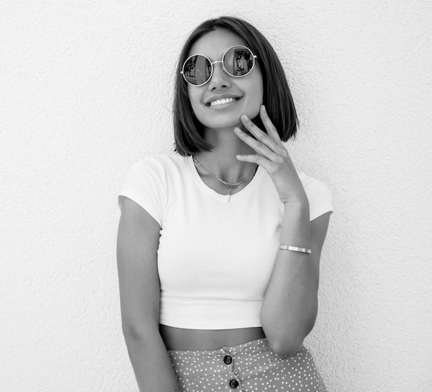 Portrait of beautiful smiling brunette model dressed in summer hipster  clothes. Trendy girl posing in the street near white wall. Funny and positive woman having fun