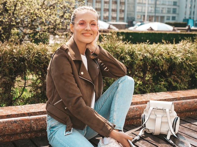 Portrait of beautiful smiling brunette model dressed in summer hipster jacket and jeans clothes  Trendy girl sitting on the bench in the street   Funny and positive woman in sunglasses