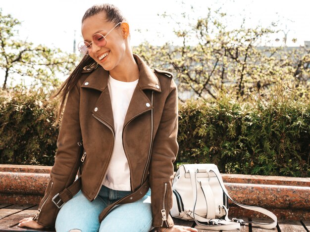 Portrait of beautiful smiling brunette model dressed in summer hipster jacket and jeans clothes  Trendy girl sitting on the bench in the street   