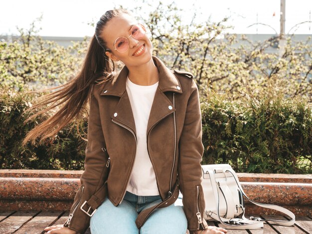 Portrait of beautiful smiling brunette model dressed in summer hipster jacket and jeans clothes  Trendy girl sitting on the bench in the street   