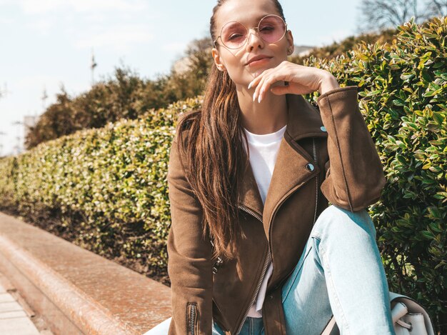 Portrait of beautiful smiling brunette model dressed in summer hipster jacket and jeans clothes  Trendy girl sitting on the bench in the street   