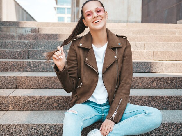 Portrait of beautiful smiling brunette model dressed in summer hipster jacket and jeans clothes. Trendy girl sitting on steps in the street background. Funny and positive woman in round sunglasses