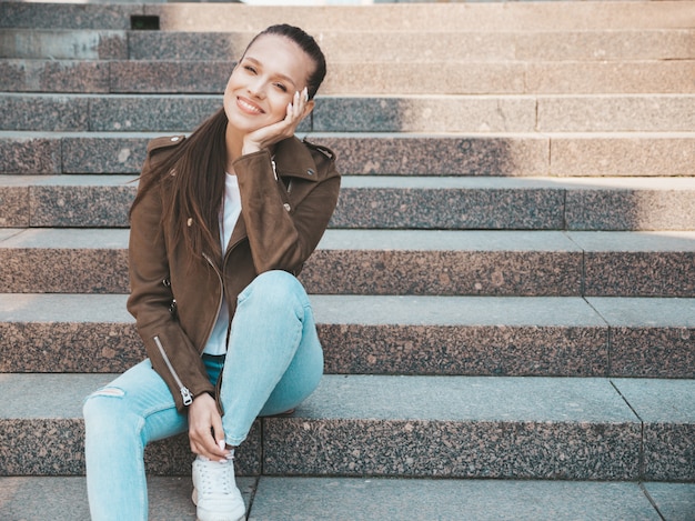 Portrait of beautiful smiling brunette model dressed in summer hipster jacket and jeans clothes. Trendy girl sitting on steps in the street background. Funny and positive woman