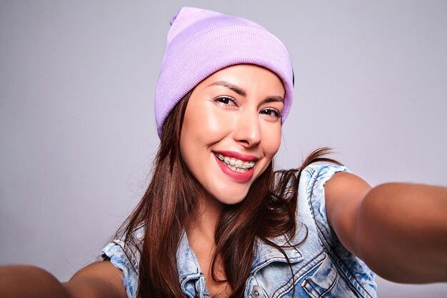 Портрет красивой улыбающейся милой брюнетки модели в повседневной джинсовой одежде без макияжа в фиолетовой шапочке, делающей селфи фото на телефон изолированную на сером