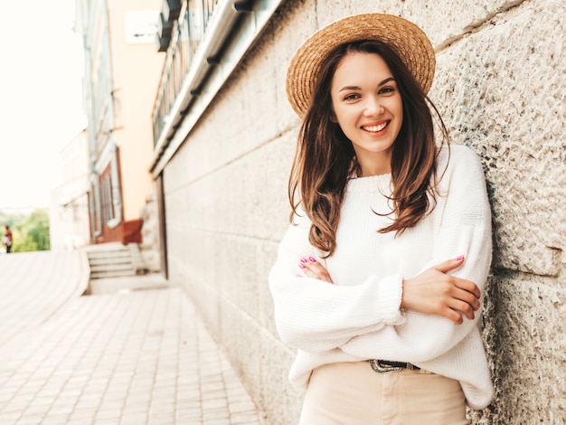 아름 다운 웃는 귀여운 모델의 초상화입니다. 따뜻한 hipster 흰색 스웨터와 모자를 입은 여성. 거리에서 벽 근처 포즈. 자신을 껴안고 재미 있고 긍정적인 여자