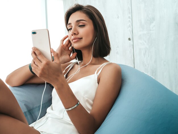 Portrait of beautiful smiling girl dressed in white pajamas and listening to music with headphones. Carefree woman sitting on soft bag chair. 