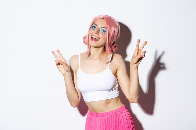 Portrait of beautiful smiling girl in pink wig, having fun on party, showing peace signs, celebrating holiday, standing.