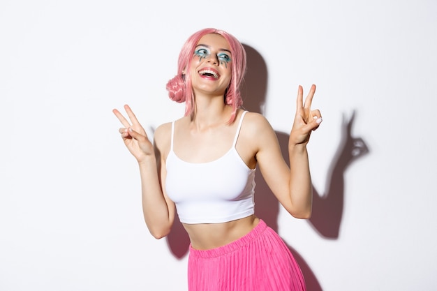 Portrait of beautiful smiling girl in pink wig, showing kawaii peace signs and laughing, wearing outfit for party, standing over white background