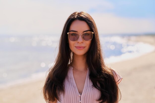 Portrait of beautiful smiling girl in sunglasses and pink dress at calm seaside.