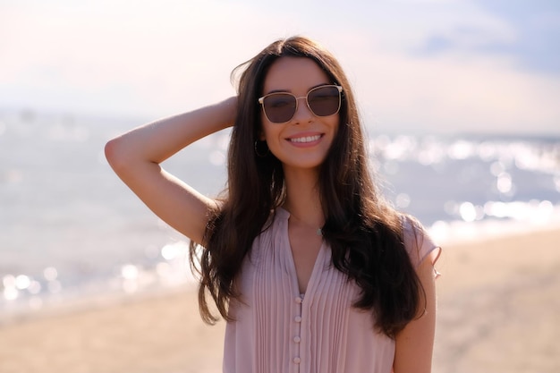 Портрет красивой улыбающейся девушки в солнечных очках и розовом платье на спокойном побережье.