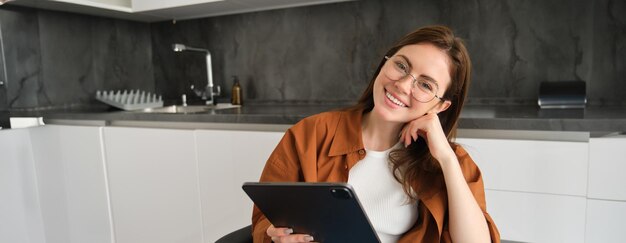 Free photo portrait of beautiful smiling girl wearing glasses sitting in kitchen with digital tablet working on