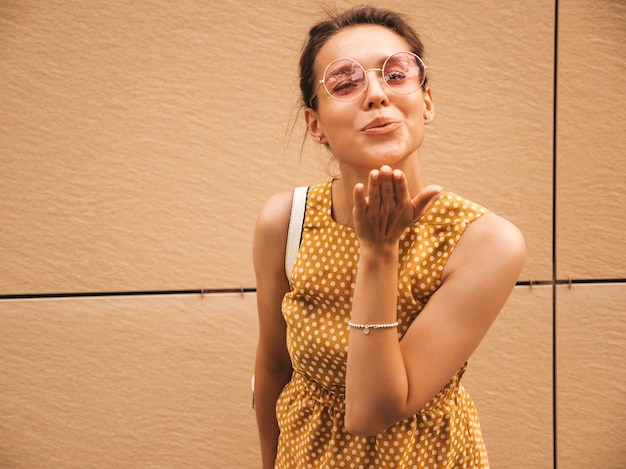 Portrait of beautiful smiling hipster model dressed in summer yellow dress. Trendy girl posing in the street. Funny and positive woman having fun.GIves air kiss