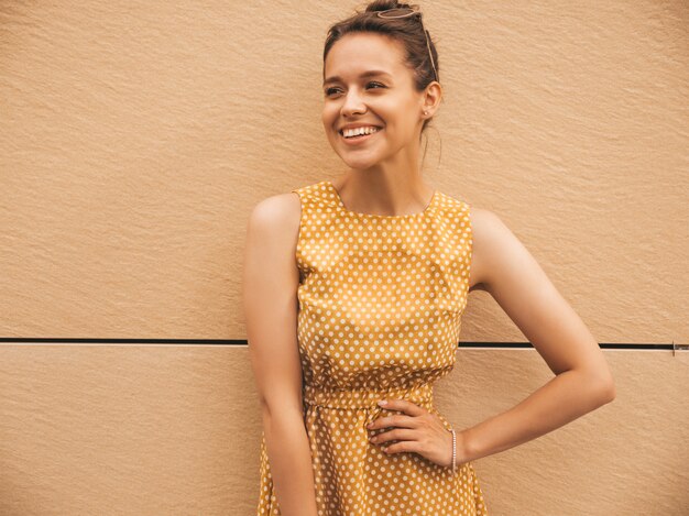 Portrait of beautiful smiling hipster model dressed in summer yellow dress. Trendy girl posing in the street. Funny and positive woman having fun