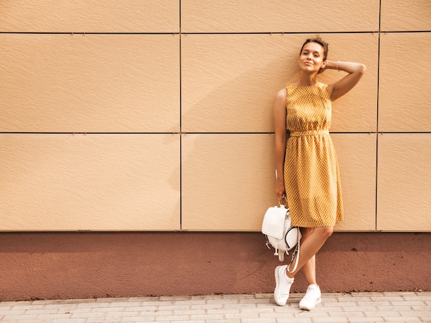 Portrait of beautiful smiling hipster model dressed in summer yellow dress. Trendy girl posing in the street. Funny and positive woman having fun