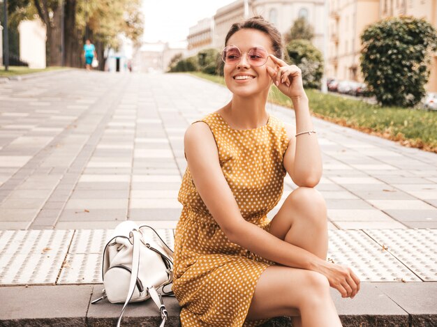 Portrait of beautiful smiling hipster model dressed in summer yellow dress. Trendy girl posing in the street. Funny and positive woman having fun