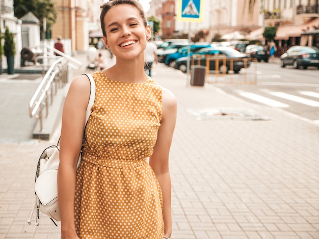 Portrait of beautiful smiling model dressed in summer yellow dress. Trendy girl posing in the street. Funny and positive woman having fun