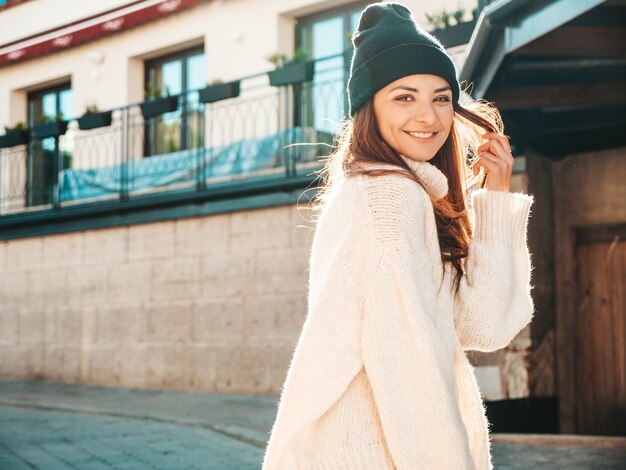 Portrait of beautiful smiling model. Female dressed in warm hipster white sweater and beanie. Posing in the street