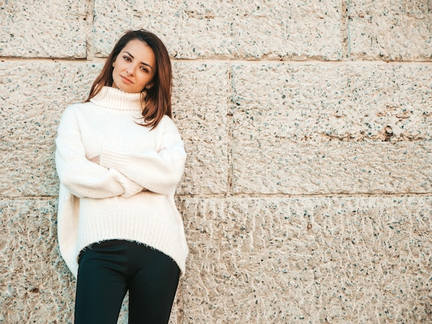 Портрет красивой улыбающейся модели. Девушка одета в теплый белый свитер битник. Позирует у стены на улице