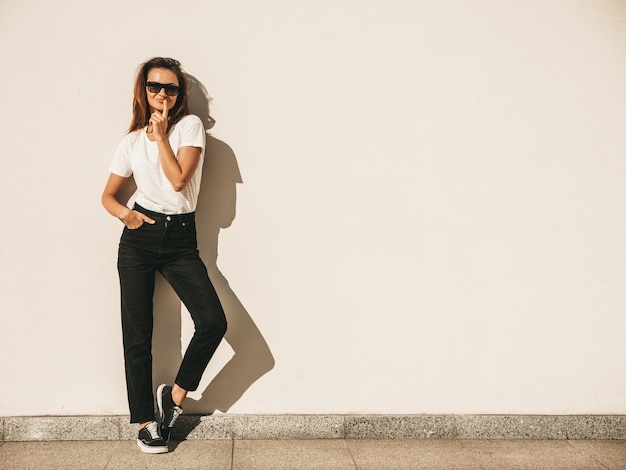 Портрет красивой улыбающейся модели в солнечных очках. Девушка, одетая в летнюю хипстерскую белую футболку и джинсы. Позирует возле стены на улице