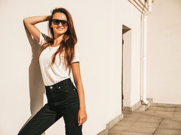 Ritratto di bella modella sorridente in occhiali da sole. donna vestita di maglietta e jeans bianchi hipster estivi. ragazza alla moda in posa vicino al muro in strada